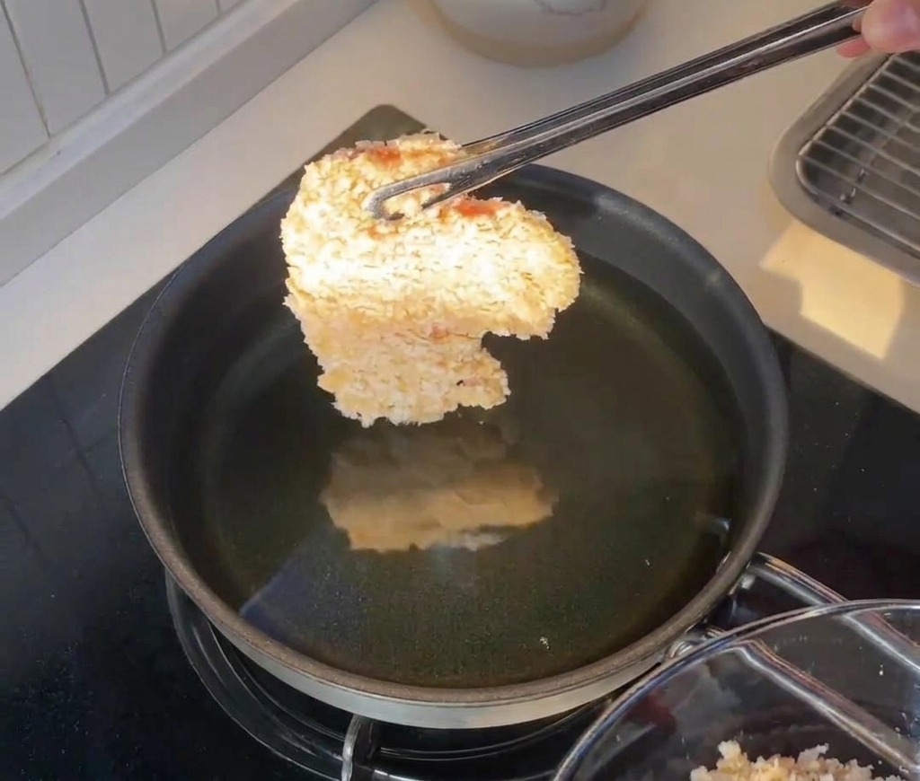 A breaded chicken fillet being held by tongs just above a frying pan filled with hot cooking oil.