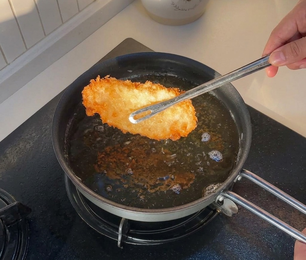 Tongs lifting a piece of golden-brown fried chicken out of a pan filled with hot oil.