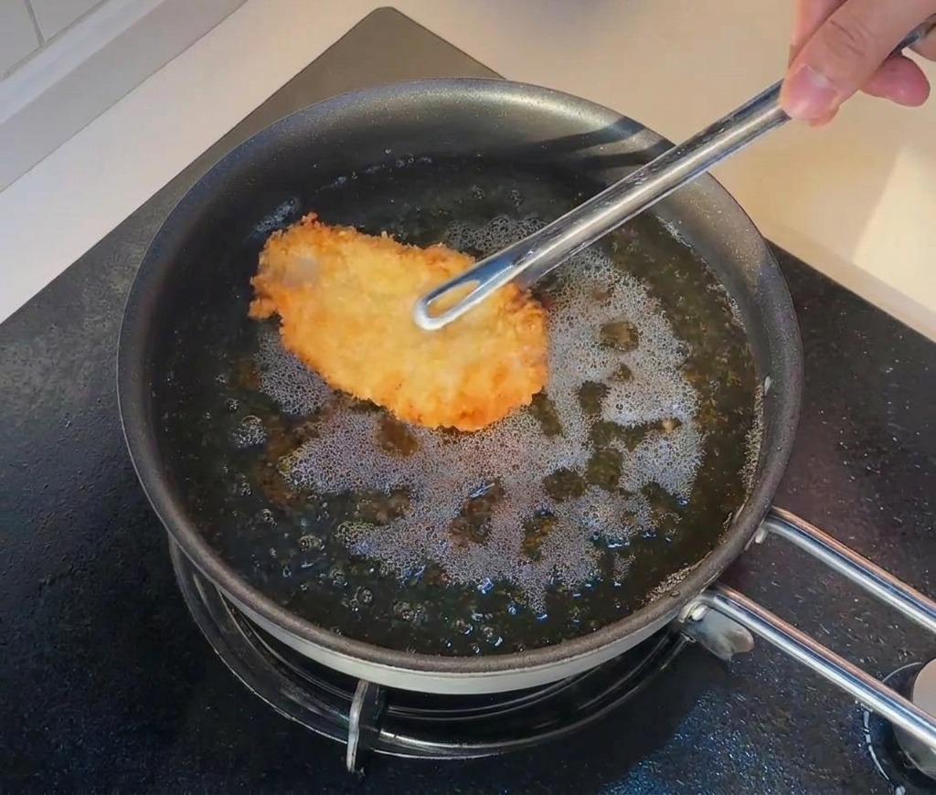 Tongs placing a piece of fried chicken back into bubbling hot oil for a second fry.