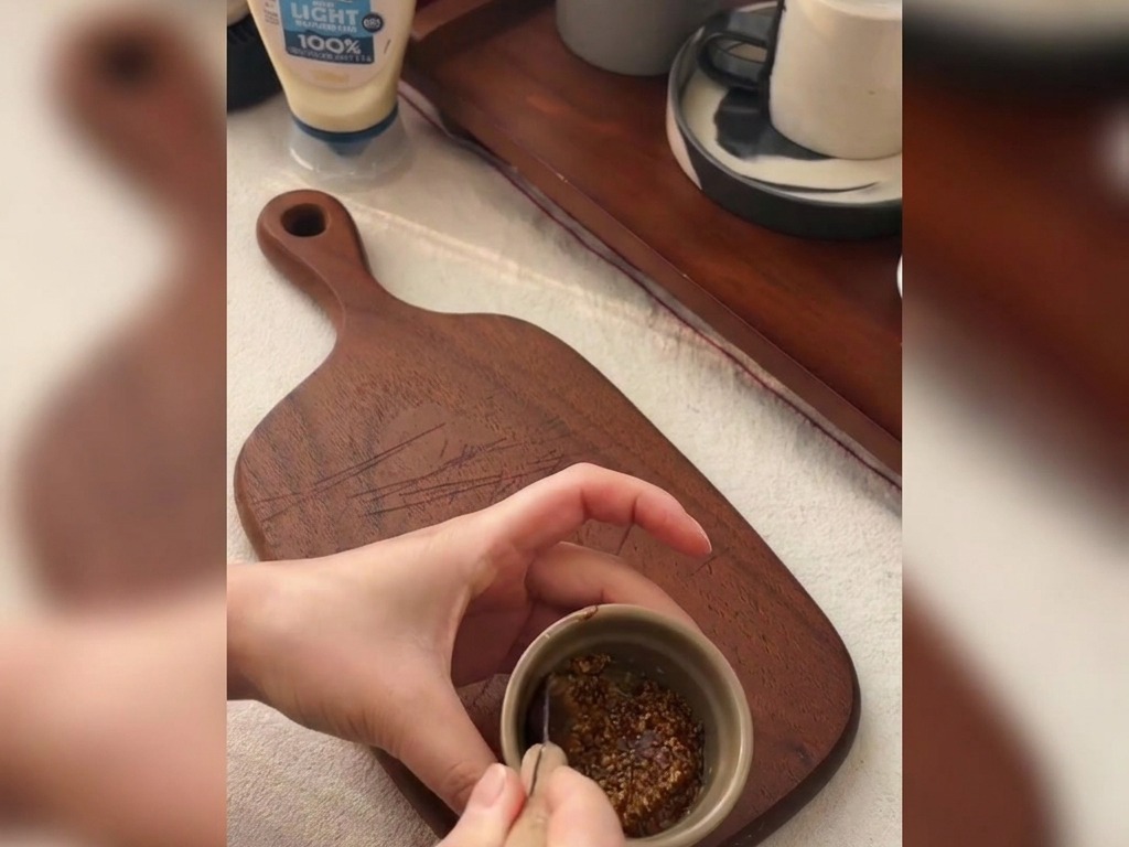 Hands using a small spoon to mix whole grain mustard and honey in a small ceramic bowl resting on a wooden cutting board.