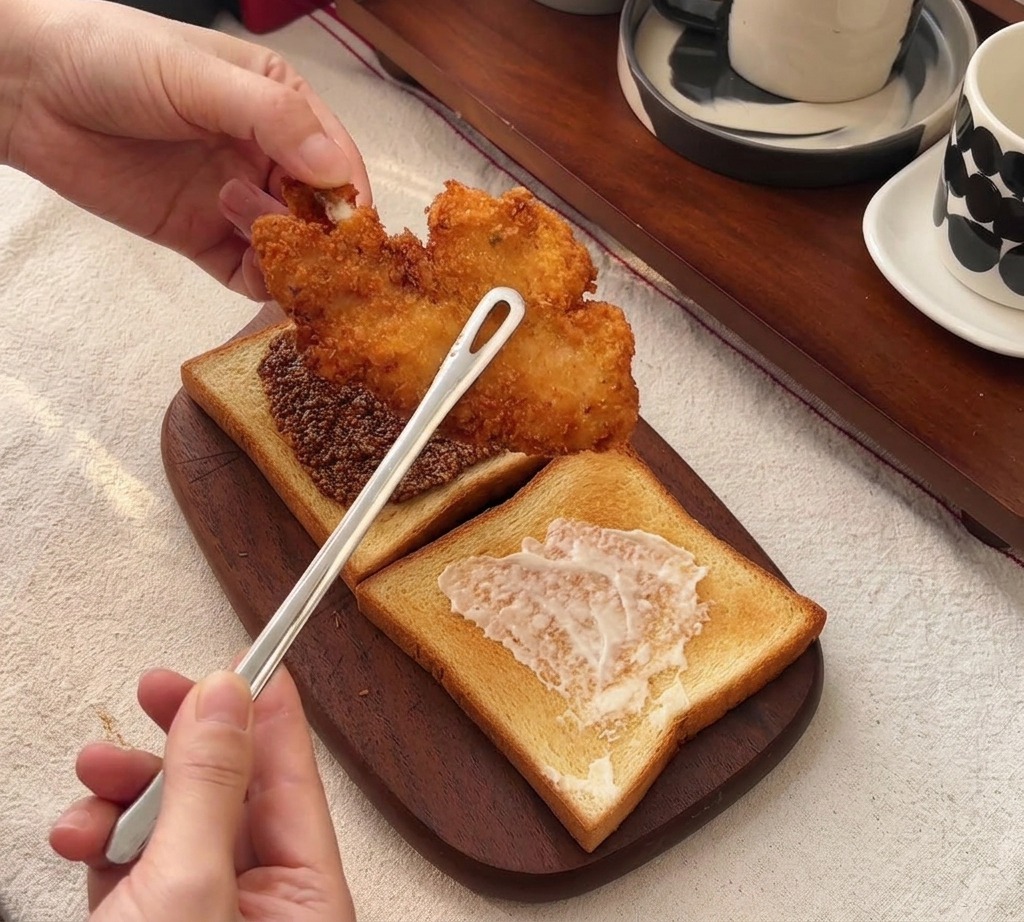 Tongs placing a large, golden-brown crispy fried chicken fillet onto a slice of toast layered with honey mustard.