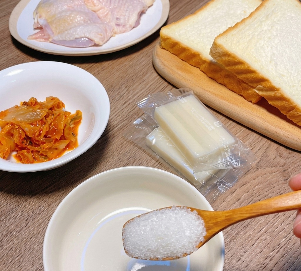 A wooden spoon holding white granulated sugar over a small white bowl, surrounded by ingredients like kimchi, bread, and raw chicken.