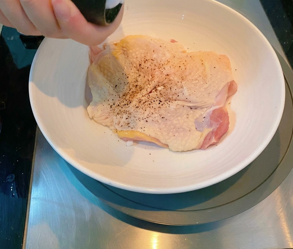 A hand using a pepper grinder to season a raw chicken thigh in a white ceramic bowl.