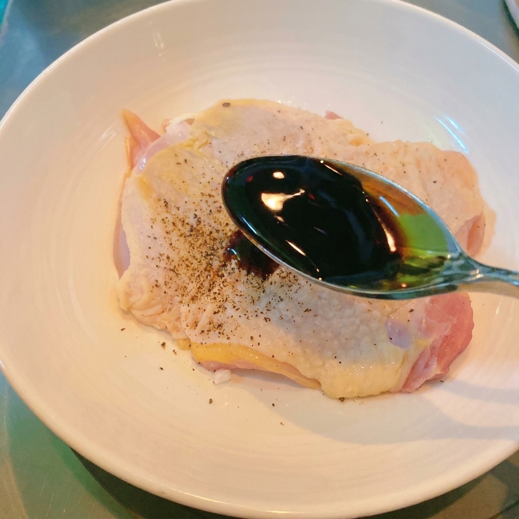 A metal spoon pouring dark soy sauce over a seasoned piece of raw chicken thigh in a white bowl.