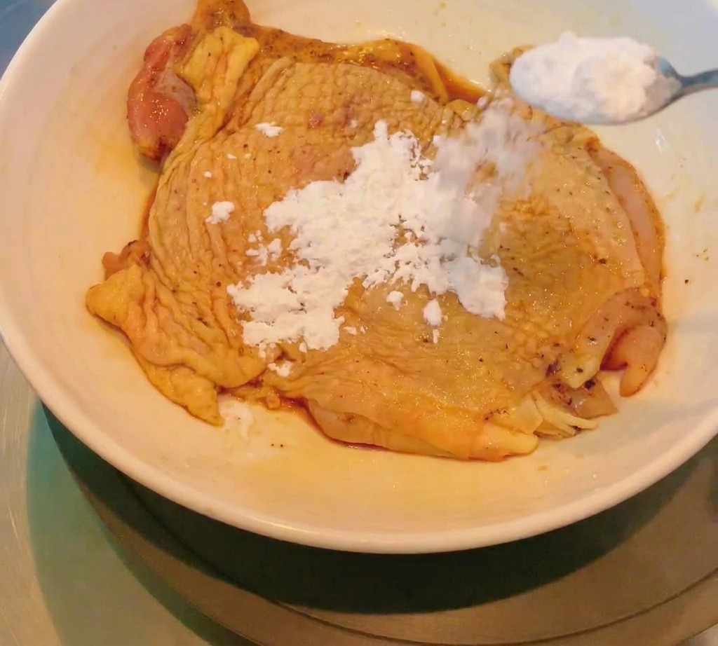 White cornstarch being spooned onto a marinated raw chicken thigh to create a thin, even coating.
