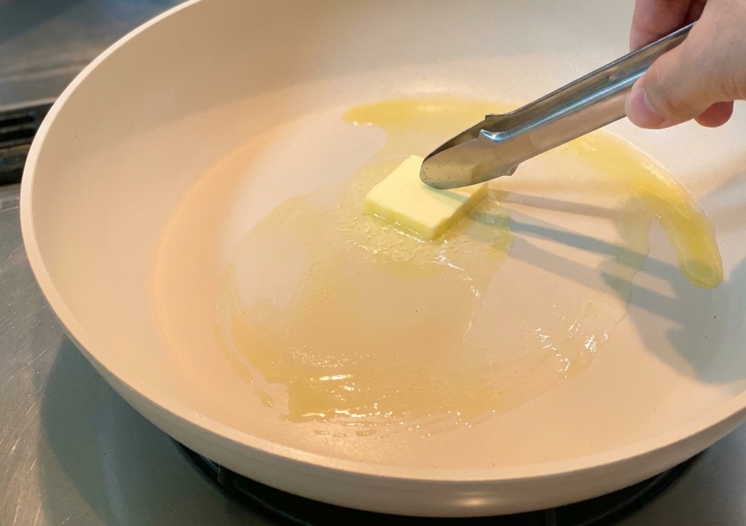 A square of butter being moved around a light-colored frying pan with metal tongs as it begins to melt.