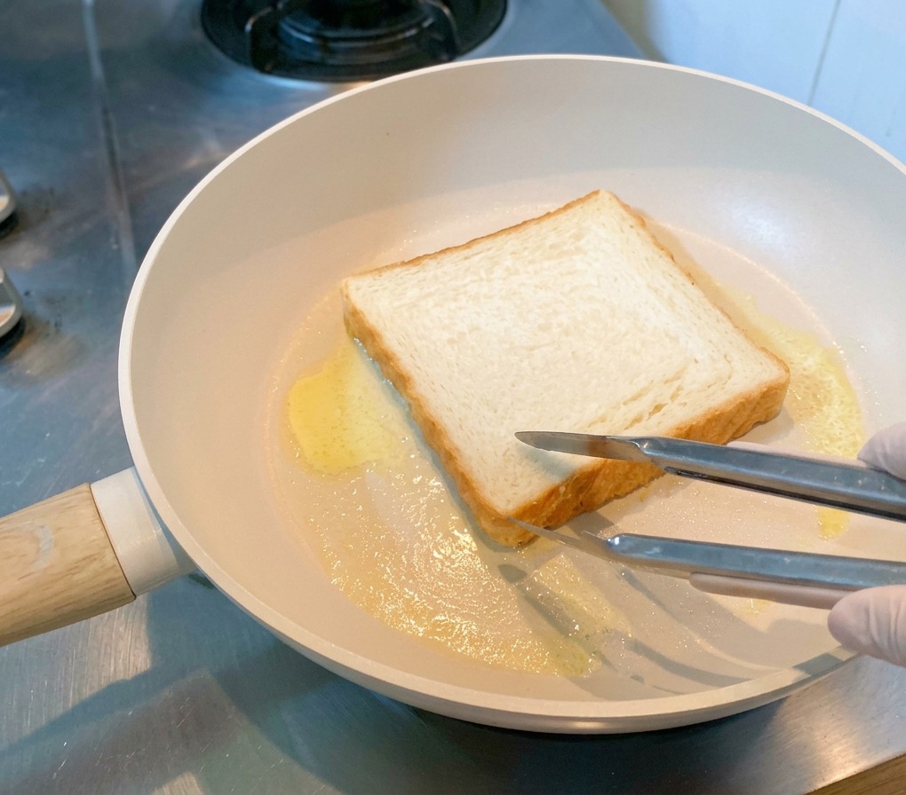 A slice of white sandwich bread being toasted in a pan with melted butter, held in place by tongs.