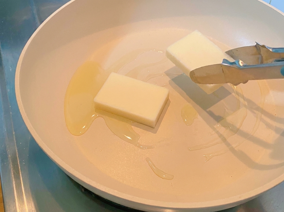 Tongs placing two thick, rectangular white rice cakes into a heated frying pan.