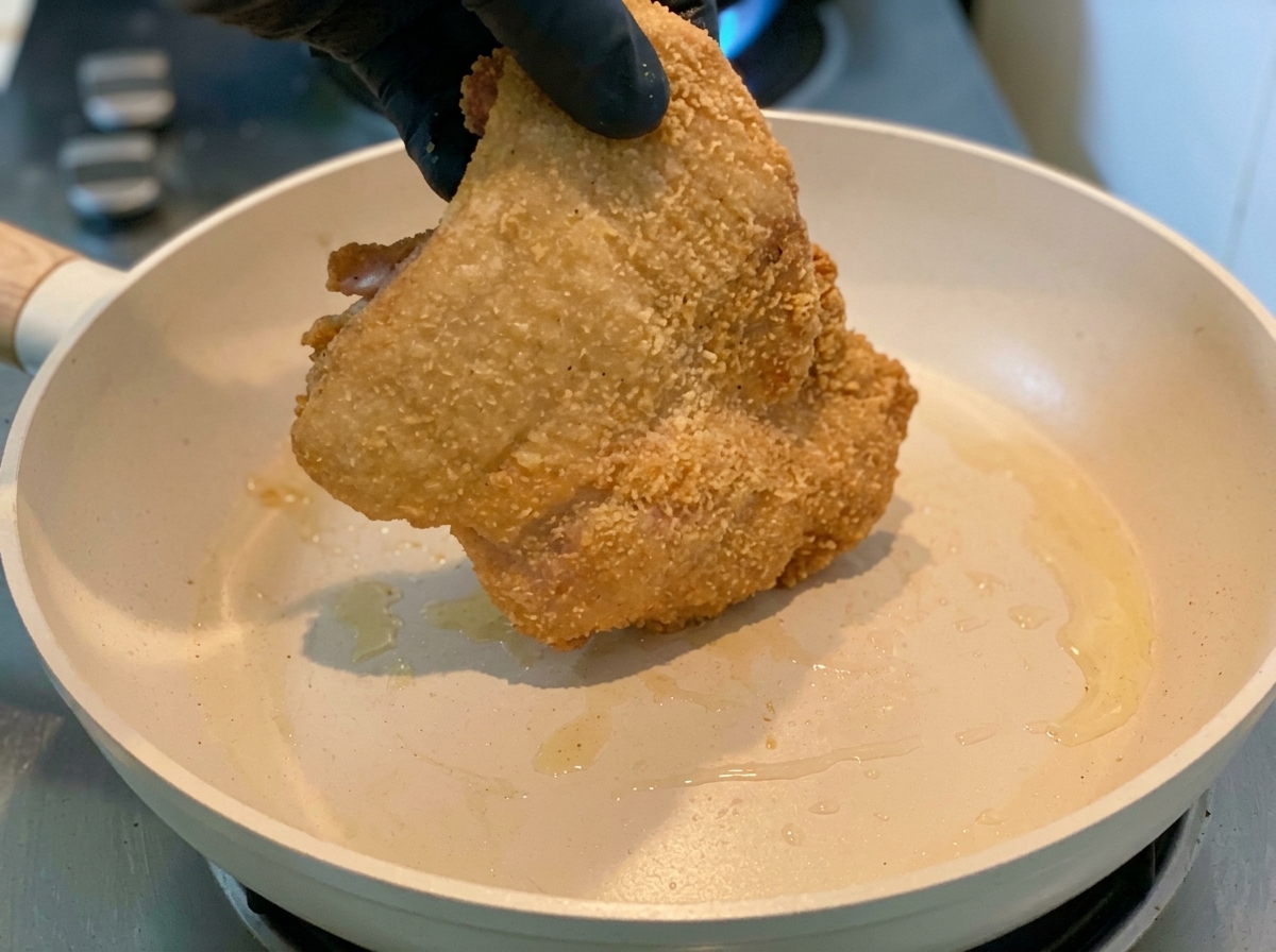 A gloved hand placing a starch coated chicken thigh into a heated frying pan.