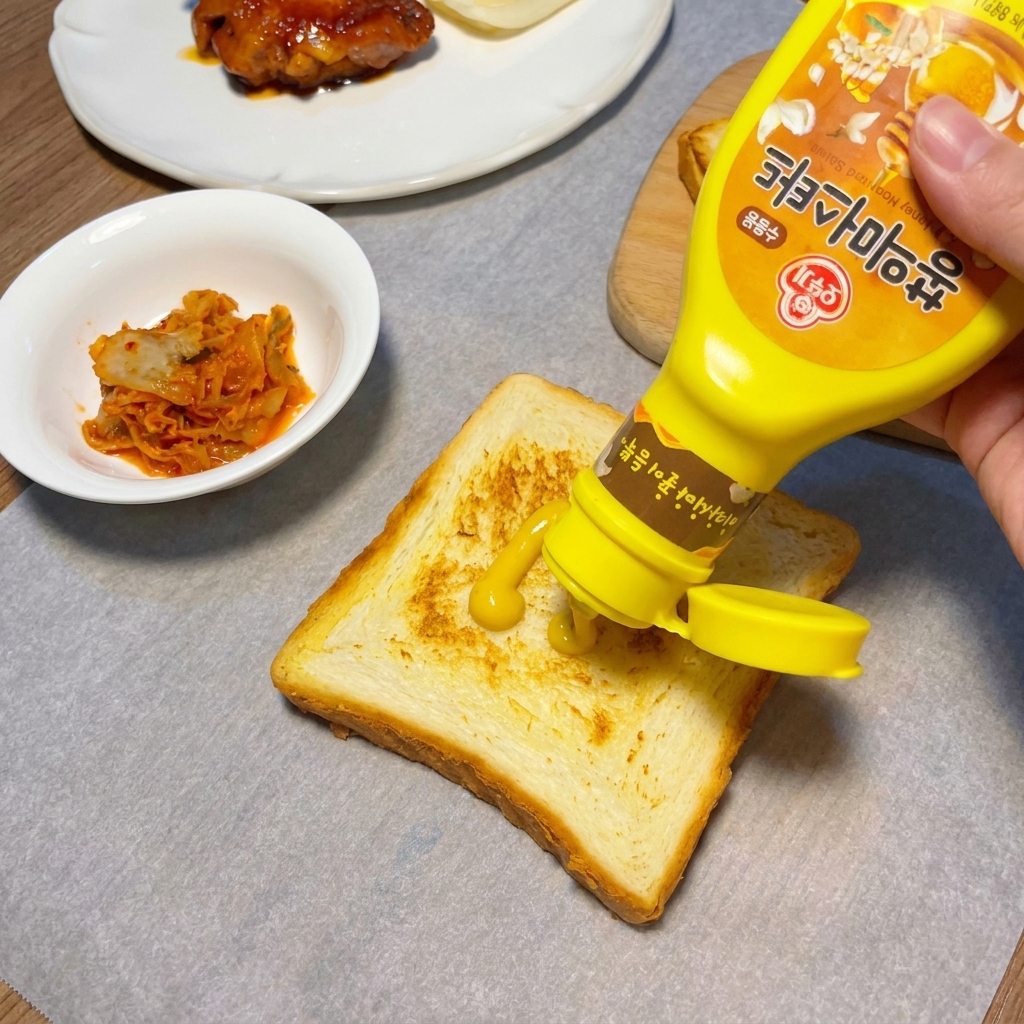 Squeezing a zigzag pattern of yellow mustard onto a golden brown slice of toasted bread.
