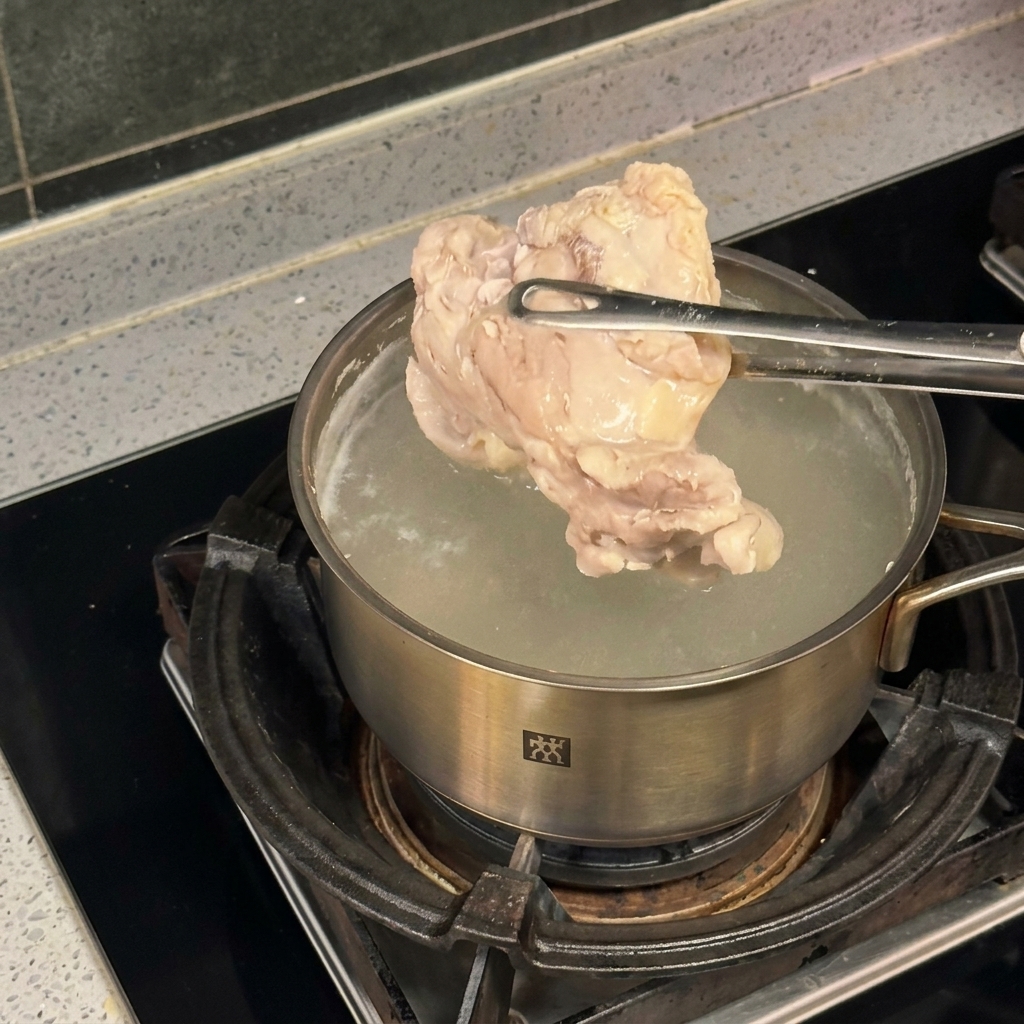A piece of chicken being held by metal tongs over a stainless steel pot of bubbling boiling water.
