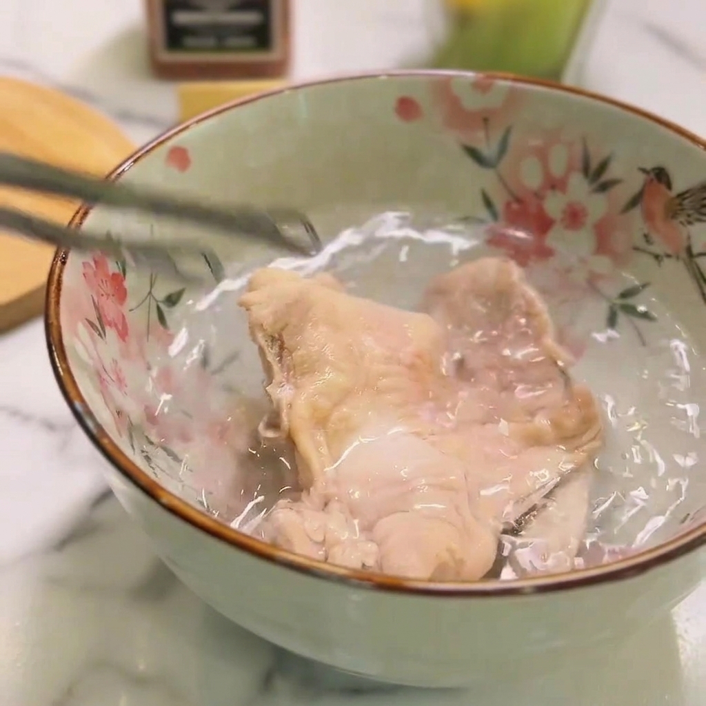 A piece of cooked chicken submerged in a floral ceramic bowl of cold water, held briefly by metal tongs.