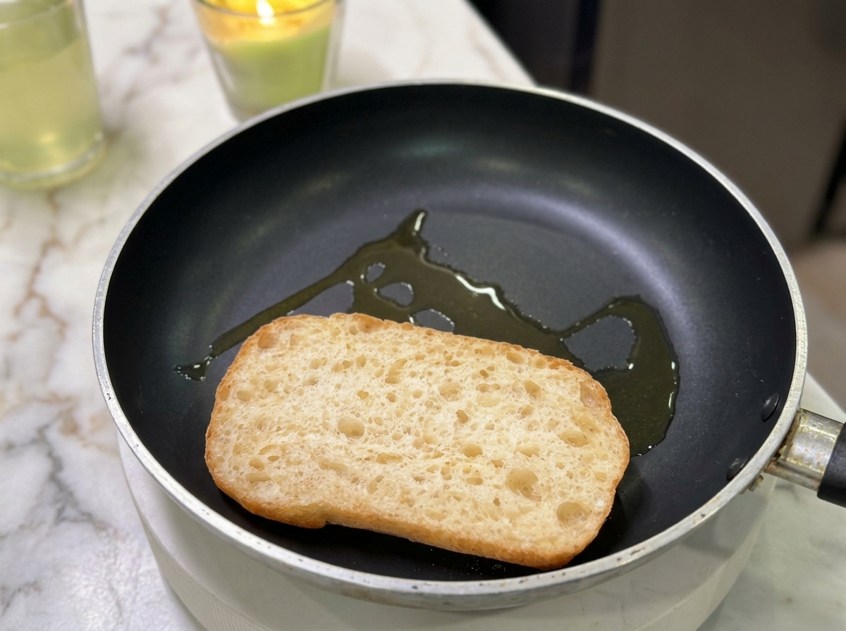 A half of a ciabatta roll toasting in a flat frying pan with a drizzle of cooking oil.