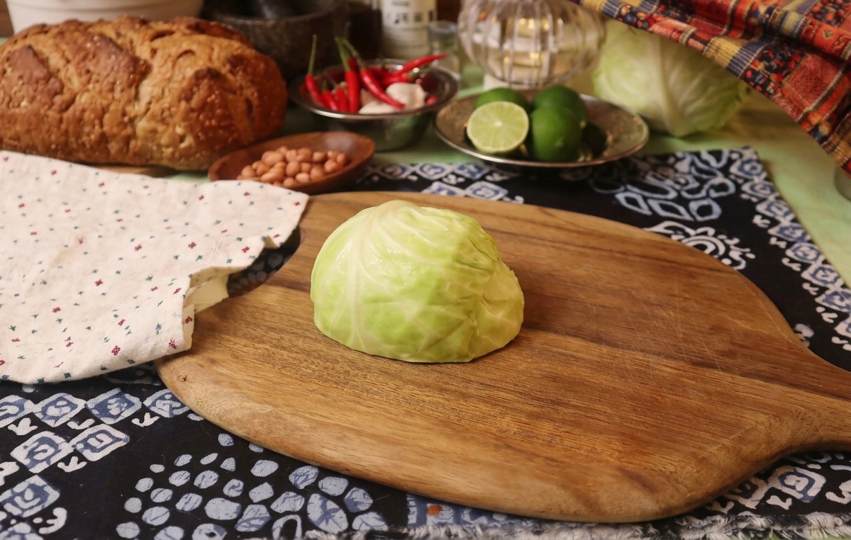 A quarter head of fresh green cabbage sitting on a wooden cutting board next to a rustic patterned cloth.