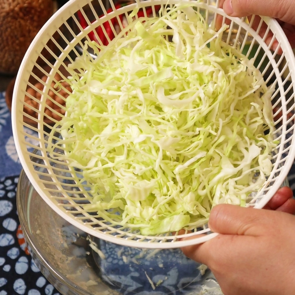 A person holding a white plastic salad spinner basket filled with freshly dried shredded cabbage.