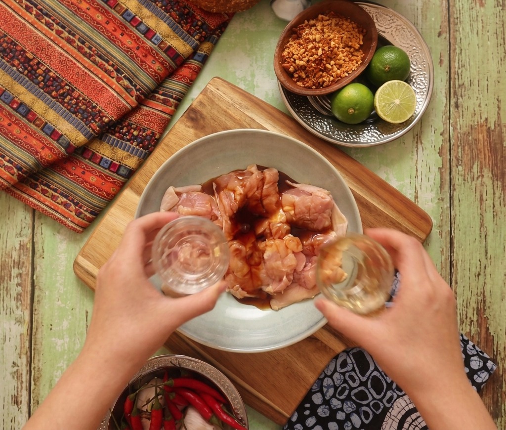 Two hands pouring liquid marinade from small glasses over raw chicken thighs in a light blue bowl.