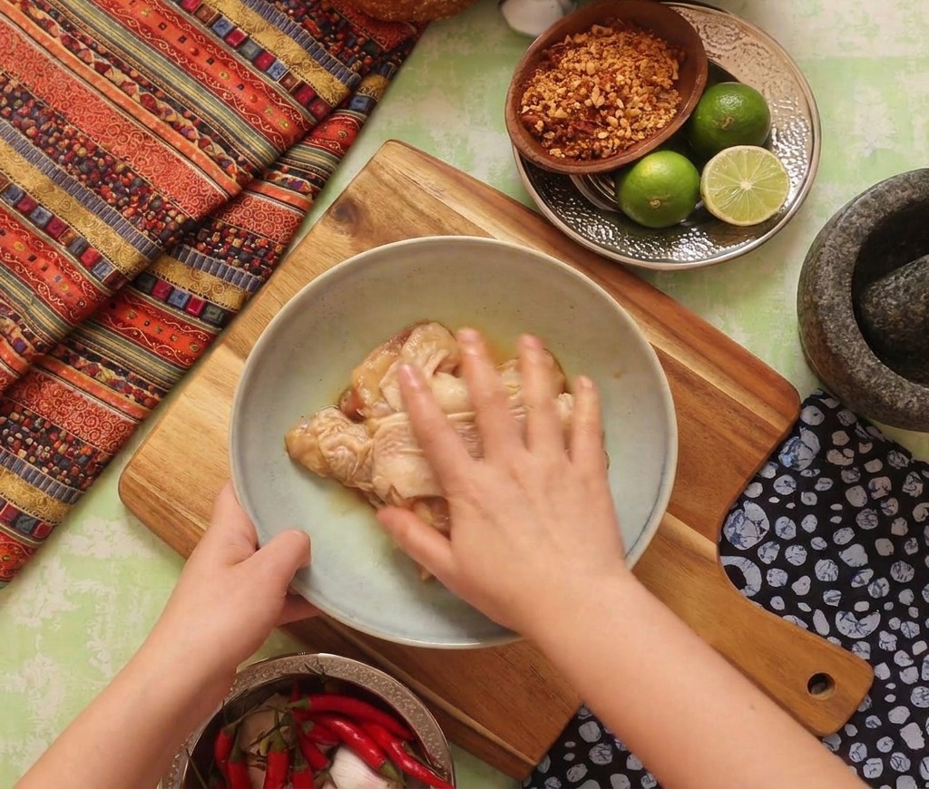 A hand mixing and massaging dark liquid marinade into raw chicken thighs in a bowl on a wooden board.