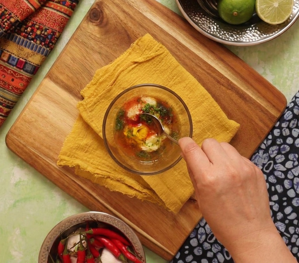 A hand stirring a bowl of creamy orange sauce made from mayonnaise, lime, and spices with a metal spoon.