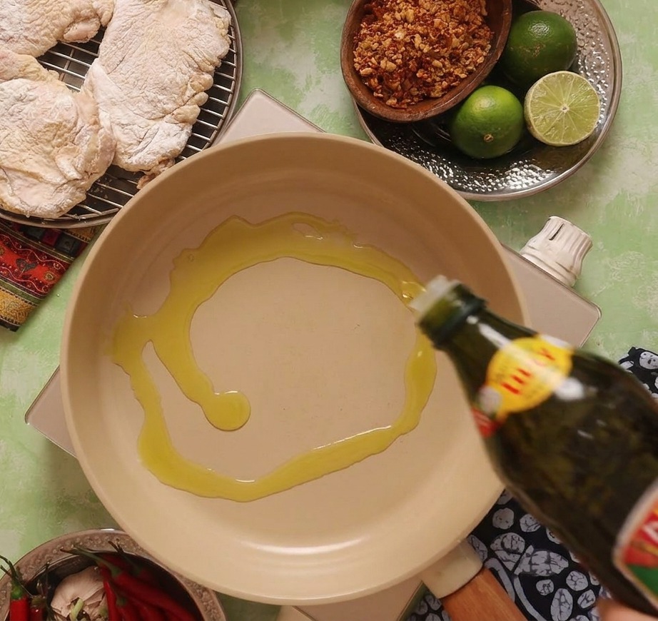 Olive oil being poured from a dark green bottle into a beige ceramic frying pan set on a portable stove.