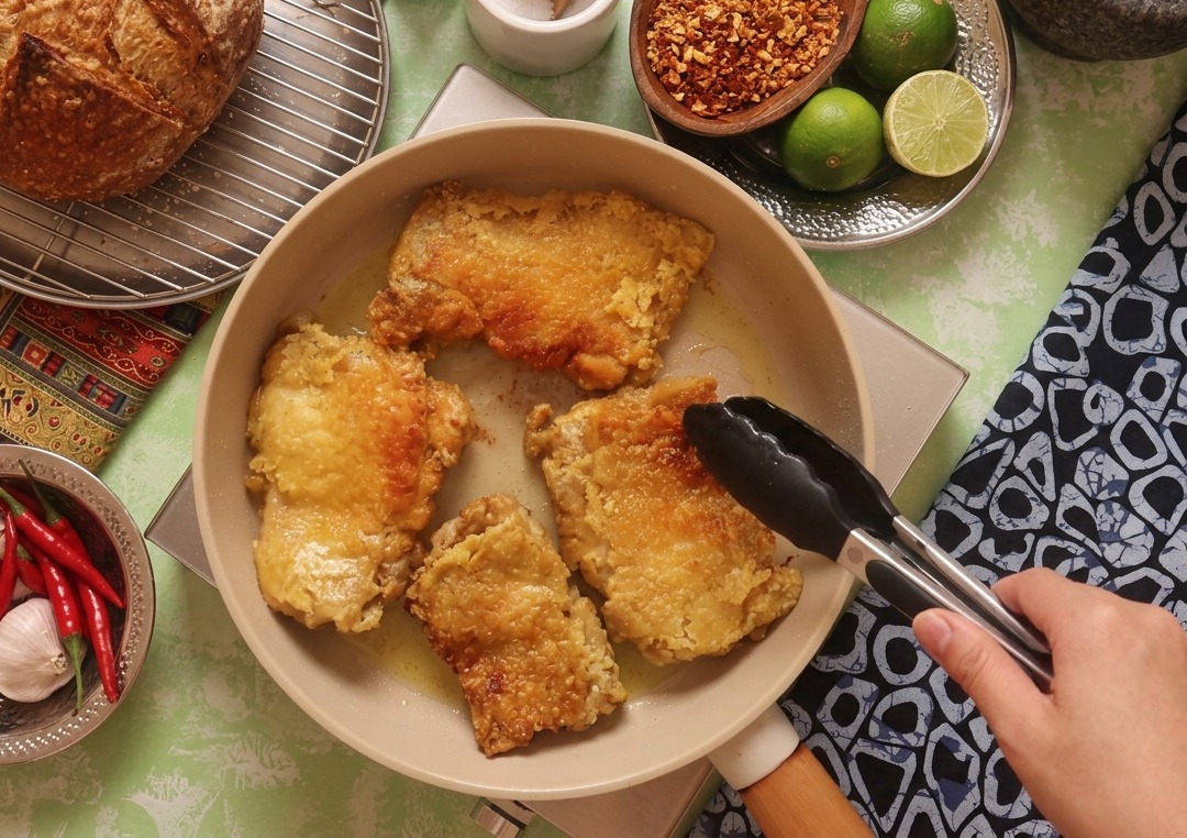 Black kitchen tongs flipping a piece of chicken in a pan, showing a perfectly golden-brown and crispy fried surface.