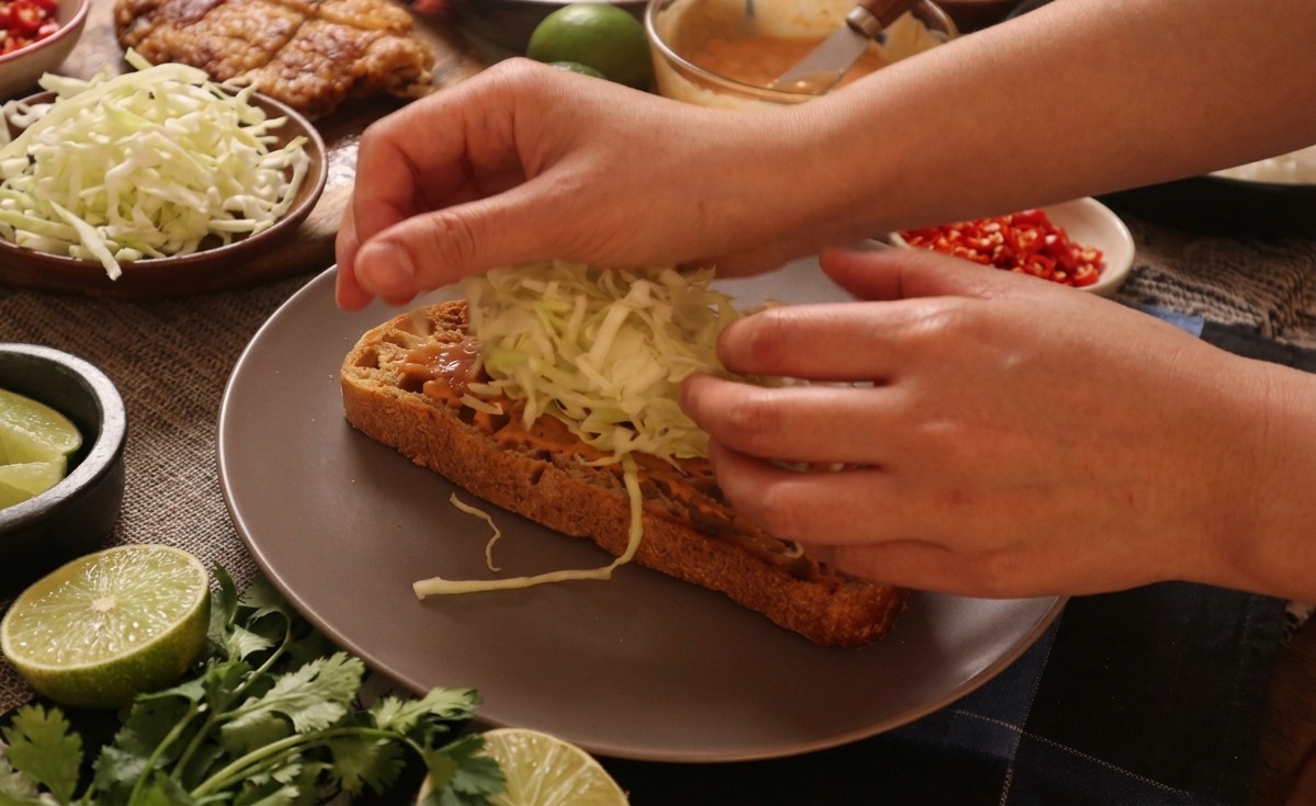 Hands placing a high mound of crisp, thinly shredded white cabbage onto a piece of sauce-topped toasted bread.