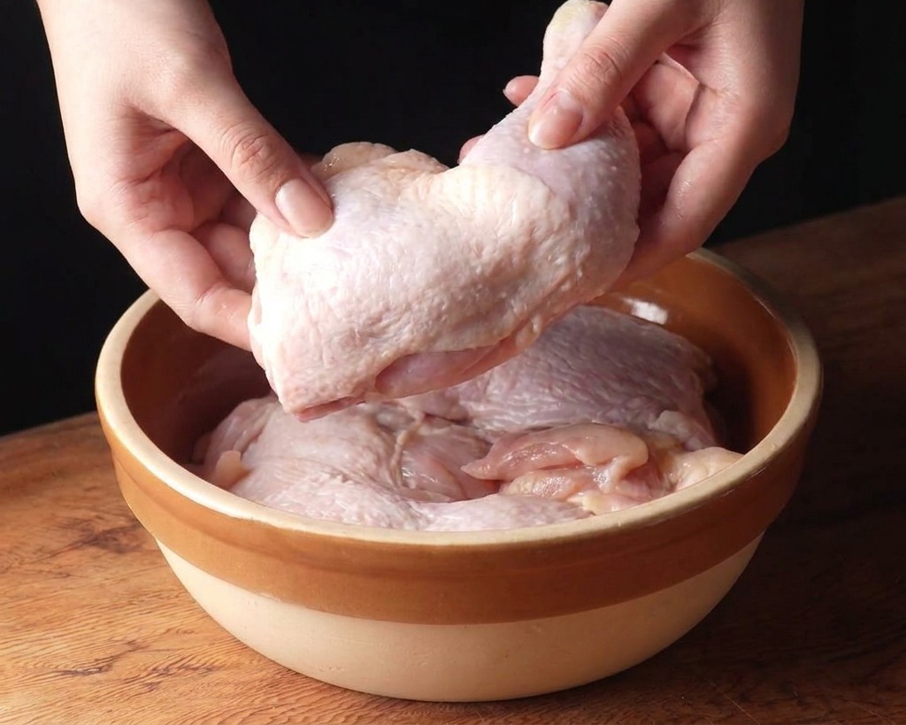 A pair of hands placing raw, skin-on chicken thighs into a beige ceramic bowl on a wooden surface.