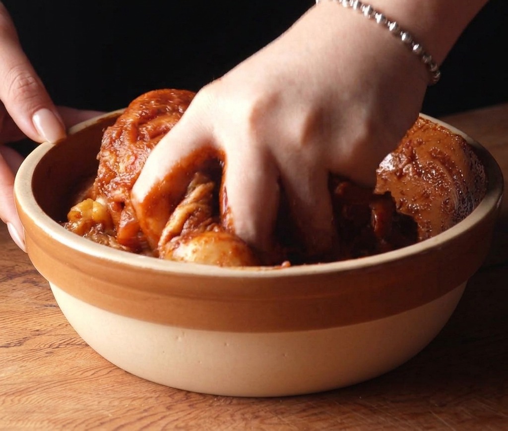 A hand mixing raw chicken thighs coated in a dark red spicy marinade inside a ceramic bowl.