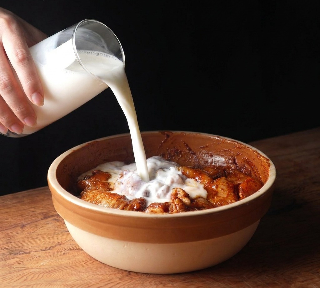 Buttermilk being poured from a glass pitcher into a ceramic bowl containing marinated raw chicken.
