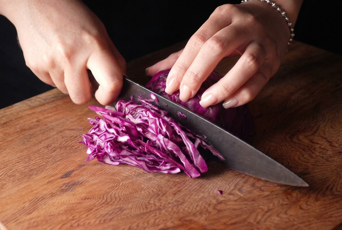 A person thinly slicing a head of purple cabbage with a chefs knife on a wooden board.