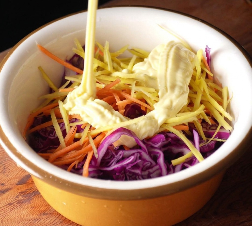 Mayonnaise being squeezed from a bottle into a bowl of shredded purple cabbage and julienned carrots.