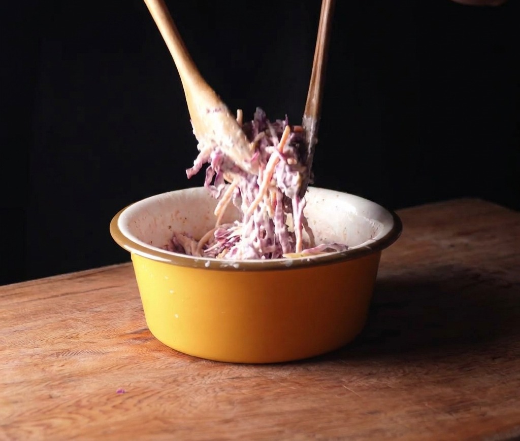 Tossing vibrant purple cabbage and carrots with creamy dressing in a yellow bowl using wooden spoons.