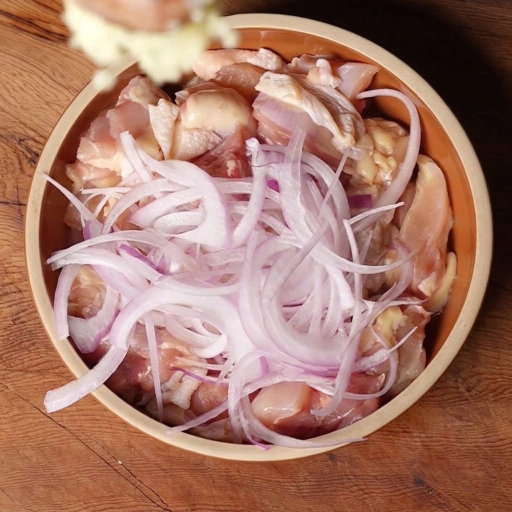 Minced garlic dropping into a ceramic bowl filled with chunks of raw chicken thigh and sliced red onions.