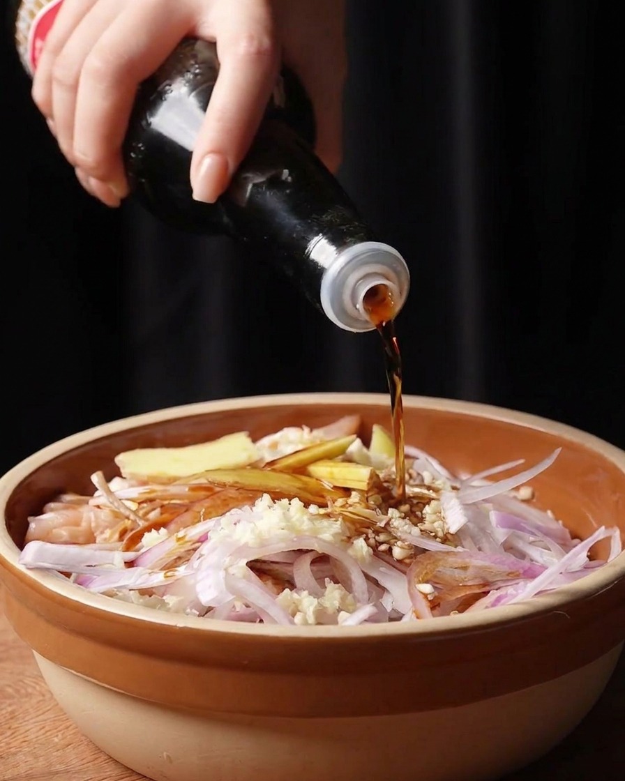 A hand pouring dark soy sauce and fish sauce from a bottle over raw chicken pieces, ginger slices, minced garlic, and red onions in a bowl.