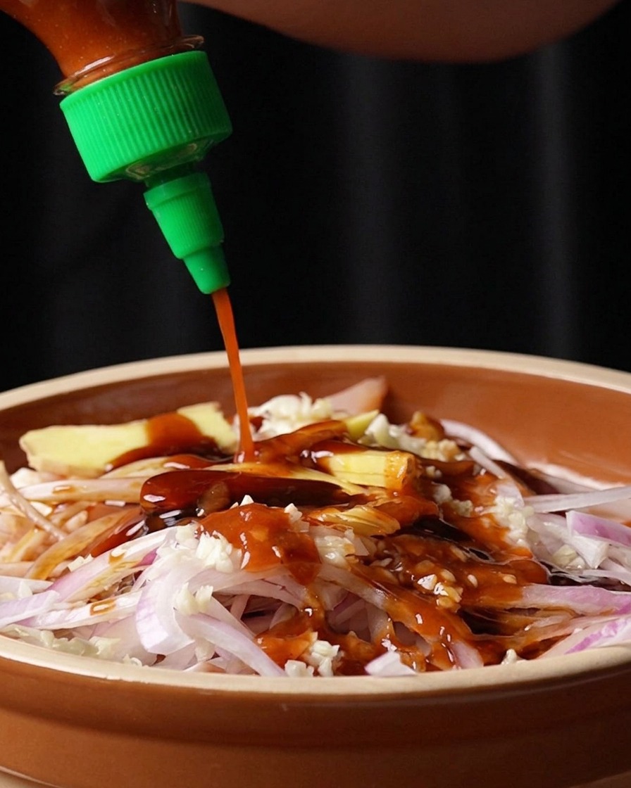 Red Sriracha chili sauce being squeezed from a green-capped bottle onto a bowl of raw chicken, onions, and aromatics.