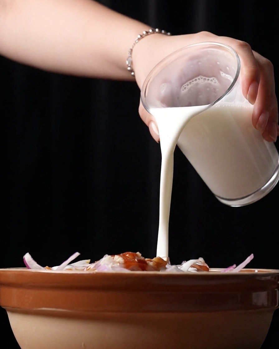 Pouring thick white buttermilk from a glass into a bowl containing raw chicken pieces and seasonings.