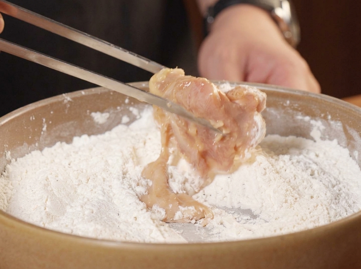 Metal tongs dropping a piece of wet marinated chicken into a dish of seasoned dry flour mixture.