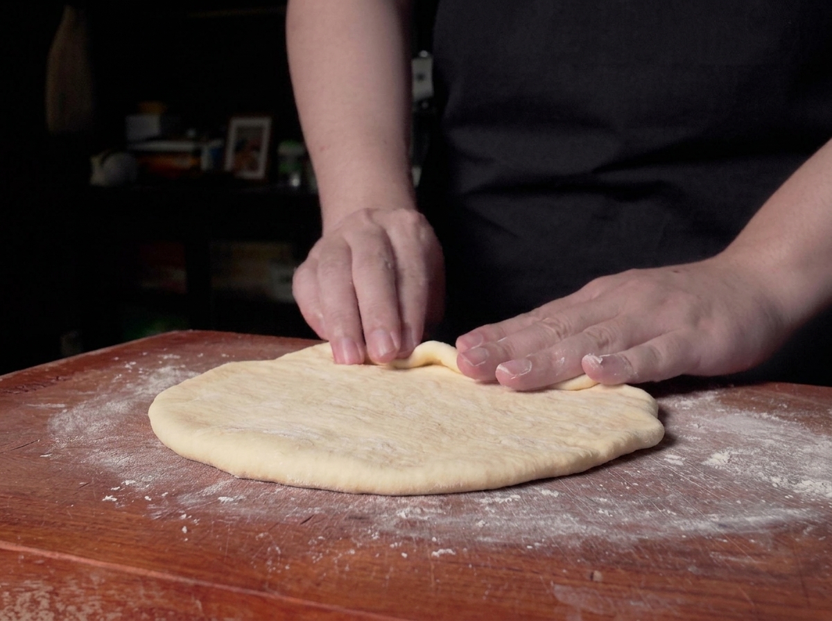 Hands beginning to roll the edge of a flat sheet of dough into a cylinder on a wooden counter.