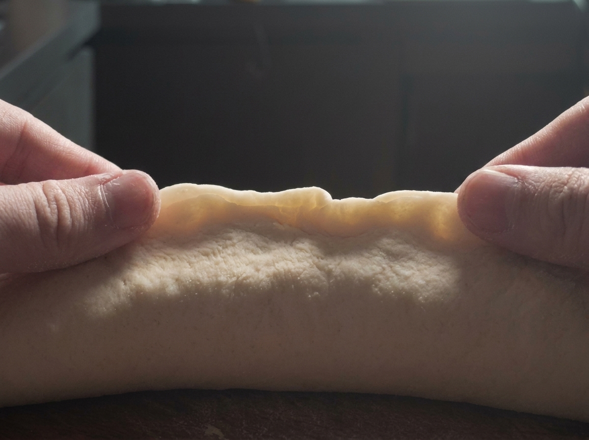 Two hands gently pinching the edge of a log of pale bread dough to seal it shut on a dark surface.