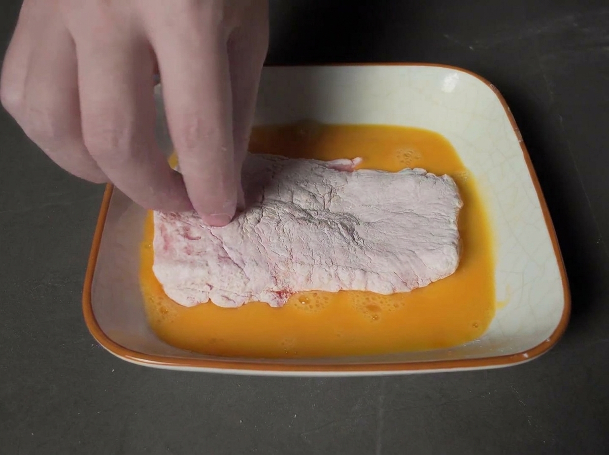 A hand dipping a flour-coated pork chop into a square dish filled with beaten golden egg wash.