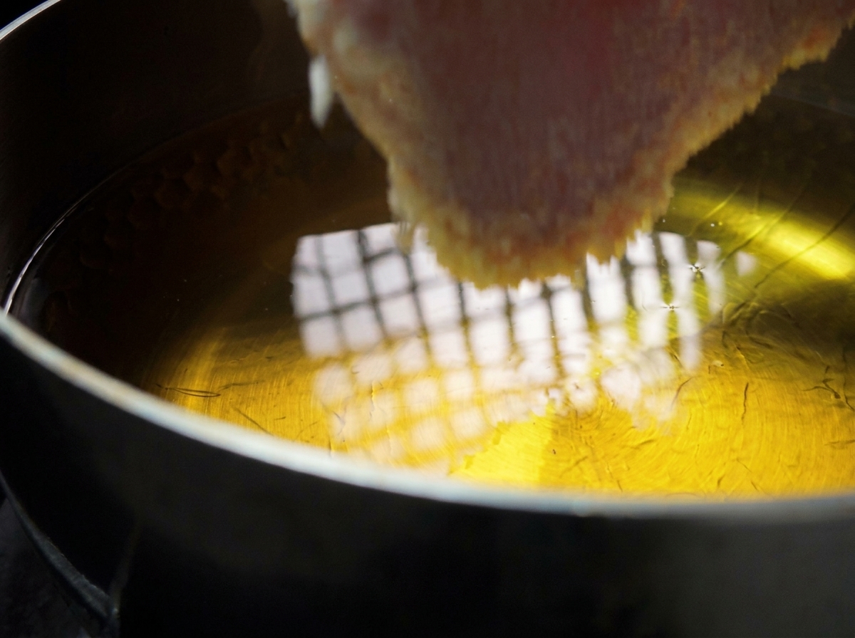 A thick, breaded pork cutlet being slowly lowered by hand into a pot of shimmering hot cooking oil.