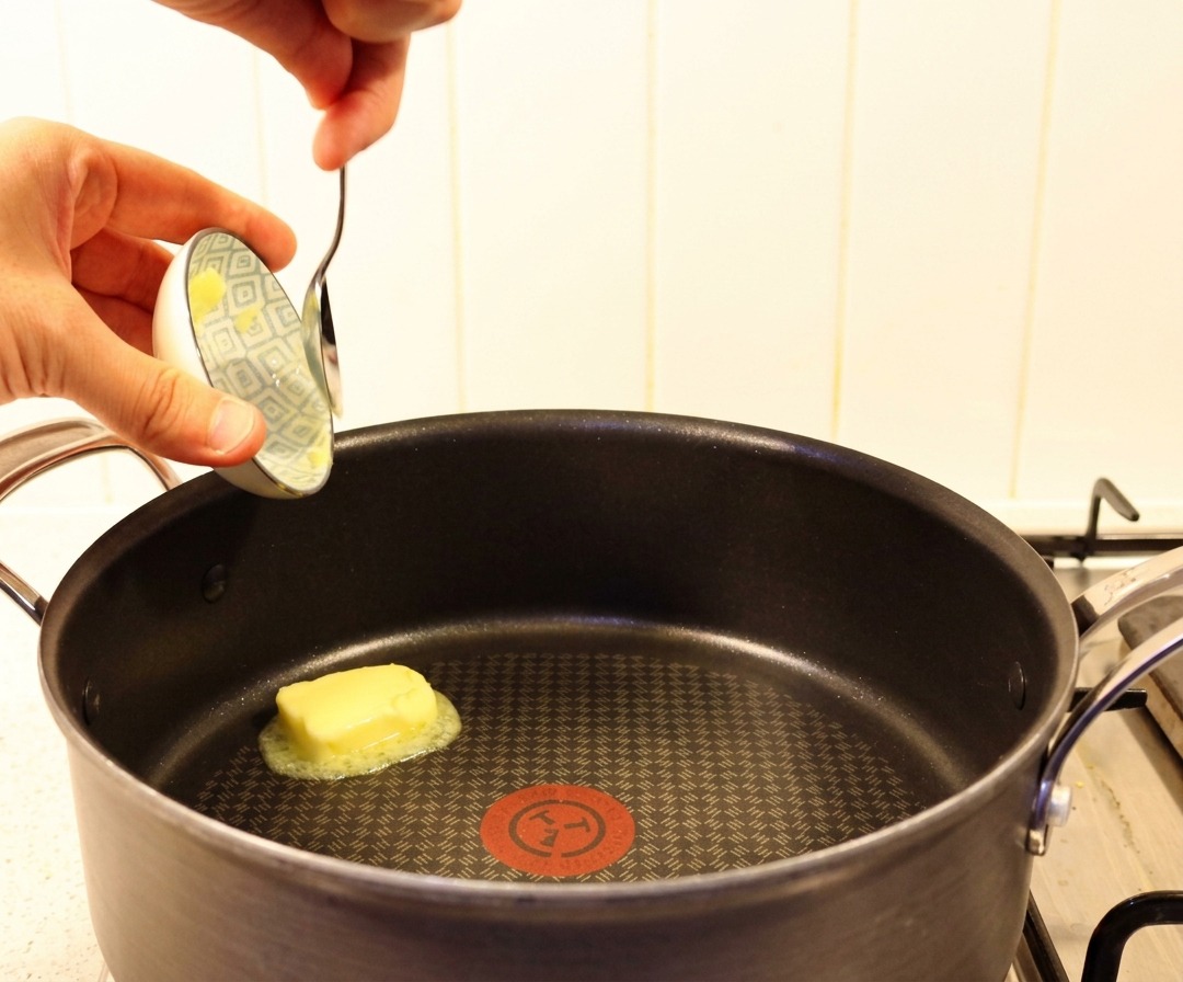 A hand using a spoon to drop a piece of yellow butter into a hot black skillet.