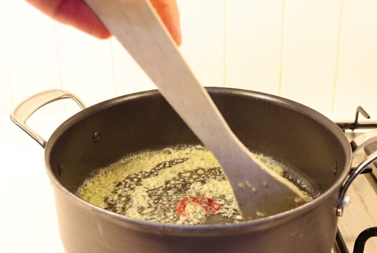 Minced garlic and chili being stirred into melted butter in a dark pot with a wooden spatula.