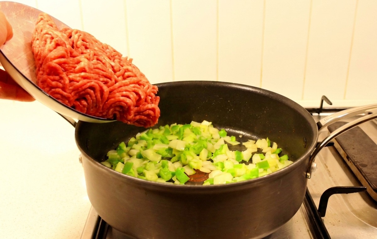 Fresh raw ground beef being poured from a plate into a skillet containing sautéed green peppers and onions.