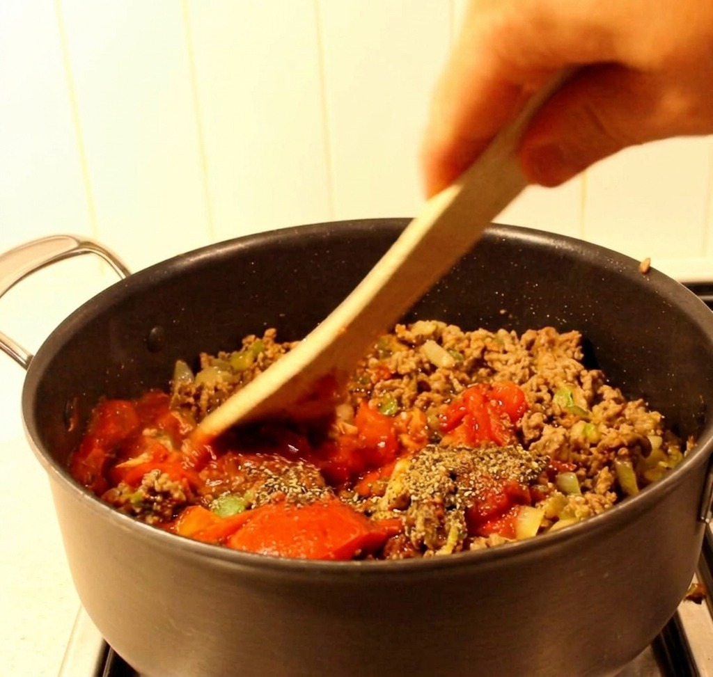 A wooden spatula mixing ground beef, green peppers, and a red tomato-based sauce together in a black saucepan.