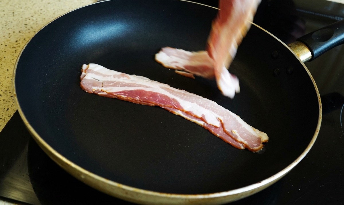 A hand placing raw bacon strips into a black frying pan.
