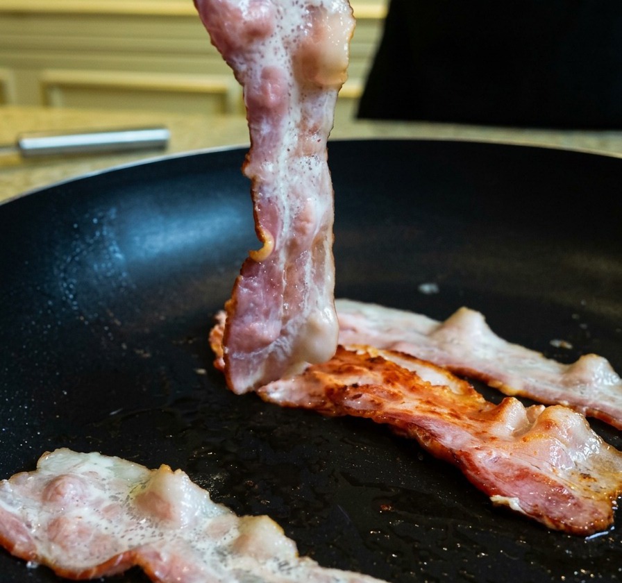 Lifting a partially cooked, browned bacon strip in a frying pan using kitchen tongs.