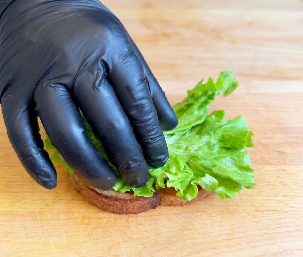 A gloved hand placing a folded piece of bright green lettuce onto a slice of toasted bread resting on a wooden board.