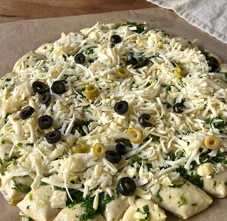 A top-down view of raw pizza dough covered in shredded cheese and topped with numerous rounds of sliced black and green olives.