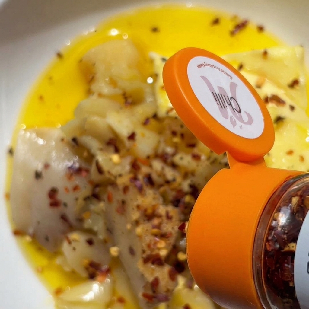 Red chili flakes being shaken from an orange-capped spice container onto a bowl of mashed garlic and butter.