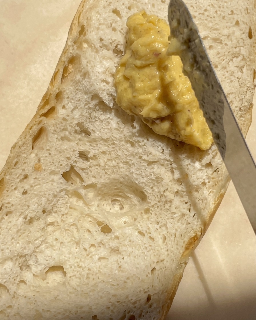 A close-up shot of a knife spreading a thick, creamy roasted garlic and butter mixture onto the airy surface of a halved baguette.